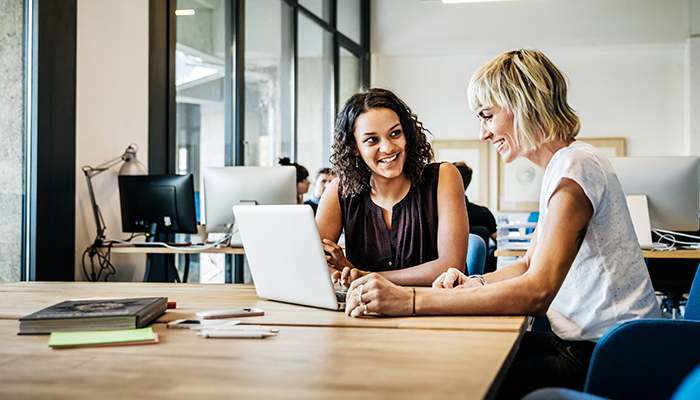 two friendly smiling people sitting at desk looking at laptop at work